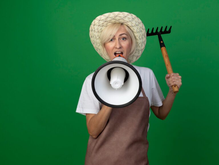 middle-aged-blonde-gardener-woman-uniform-wearing-hat-raising-up-rake-talking-by-speaker-isolated-green-wall-with-copy-space