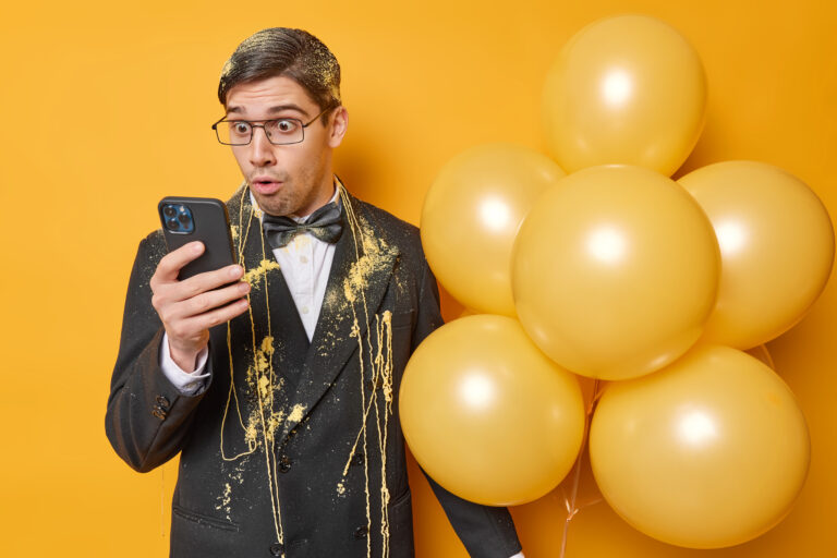 Shocked brunet man stares at smartphone screen reads awesome news dressed formally spends free time on party holds bunch of inflated balloons isolated over vivid yellow background