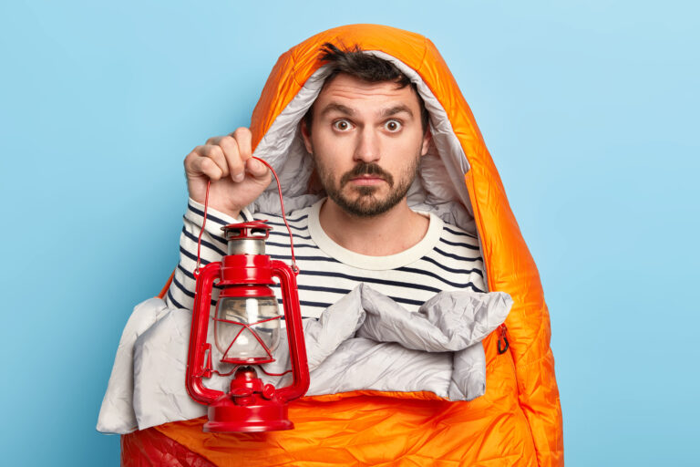 Surprised man has rest, wrapped in sleeping bag, holds kerosene lamp, enjoys recreation in forest, has expedition, poses against blue background, has adventure trip